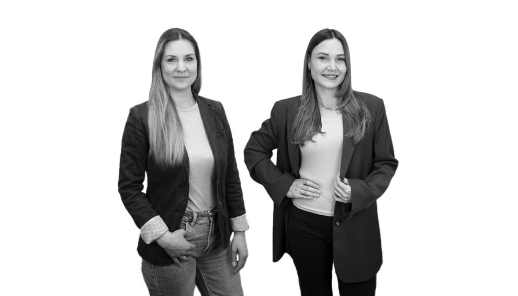 Black-and-white portrait of two women standing side by side in business attire against a white background.